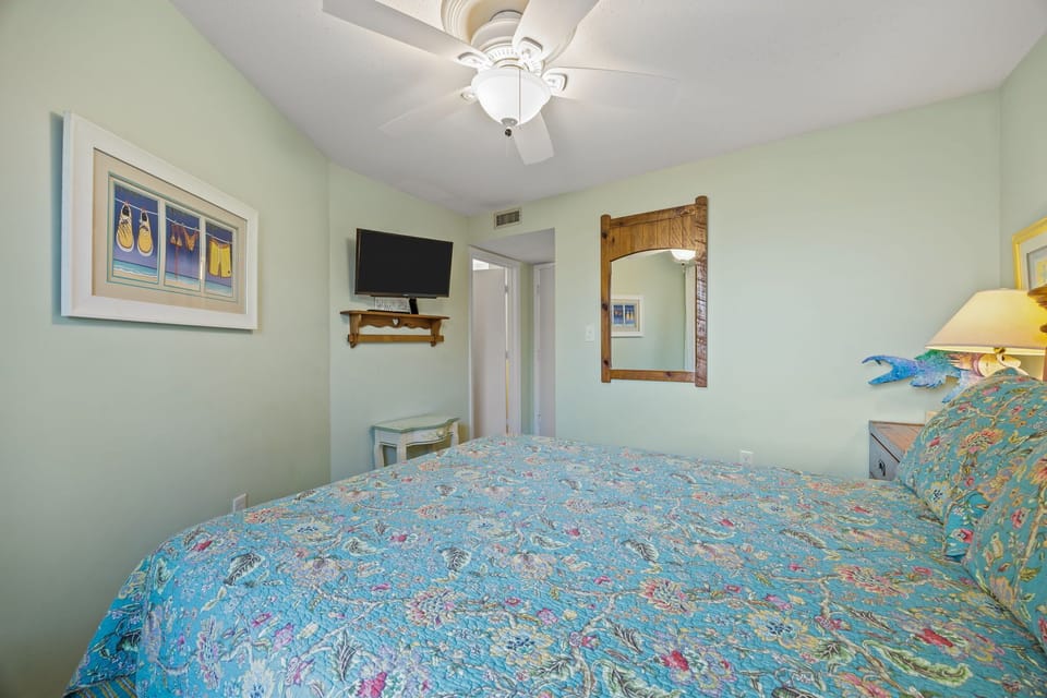Unwind in your peaceful bedroom featuring tropical-themed linens, a ceiling fan for comfort, and entertainment at your fingertips with the mounted TV.