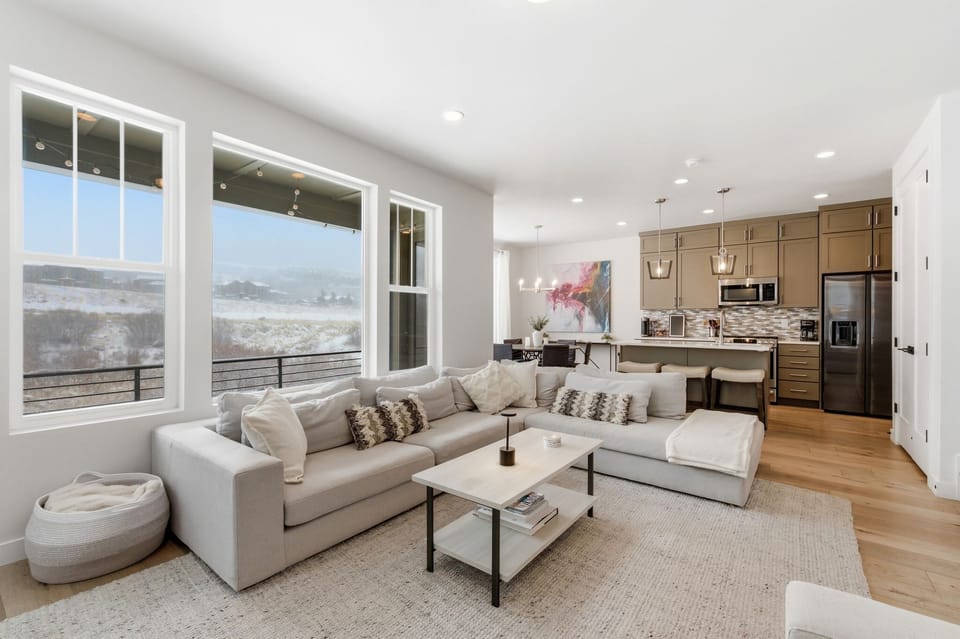 Modern living area with a comfortable sectional sofa, natural light from large windows, and an open flow into the kitchen and dining space.
