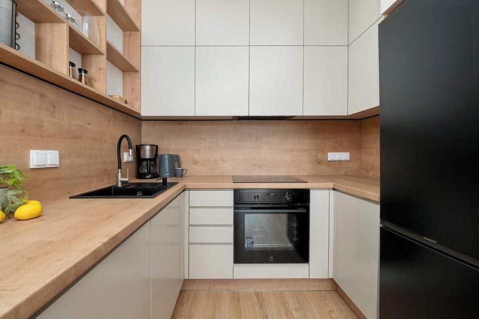 The kitchenette corner displays an L-shaped layout with clean lines and a high-contrast black refrigerator. Open wooden shelving above the counter provides space for both decor and kitchen essentials.