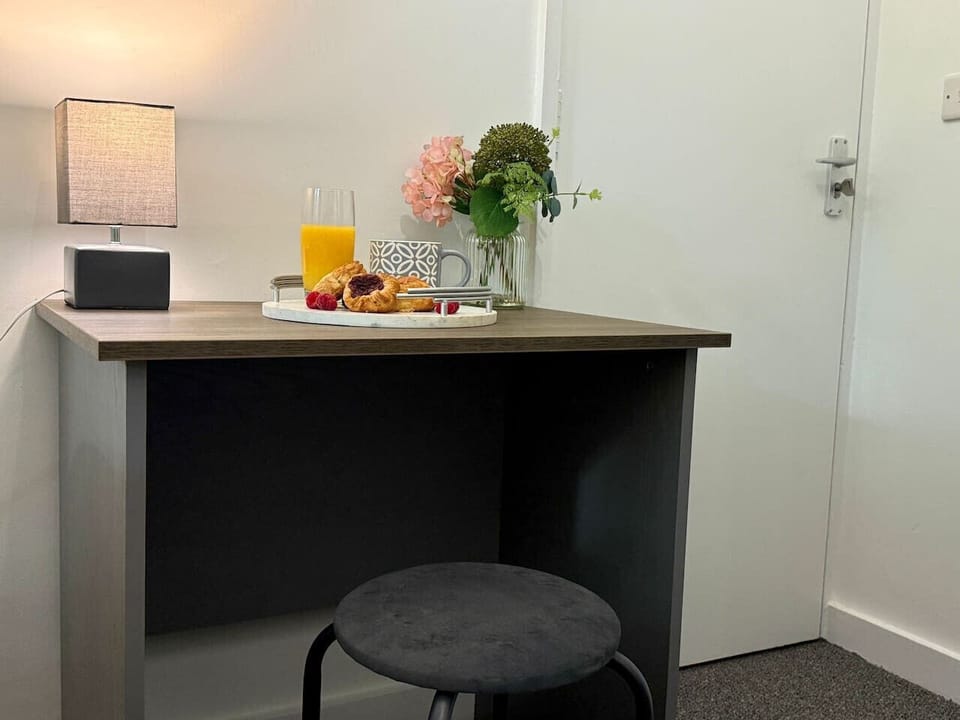 A desk area showing a lamp, a vase of pink flowers, and a glass of orange juice on a dark-topped desk with a white door behind.