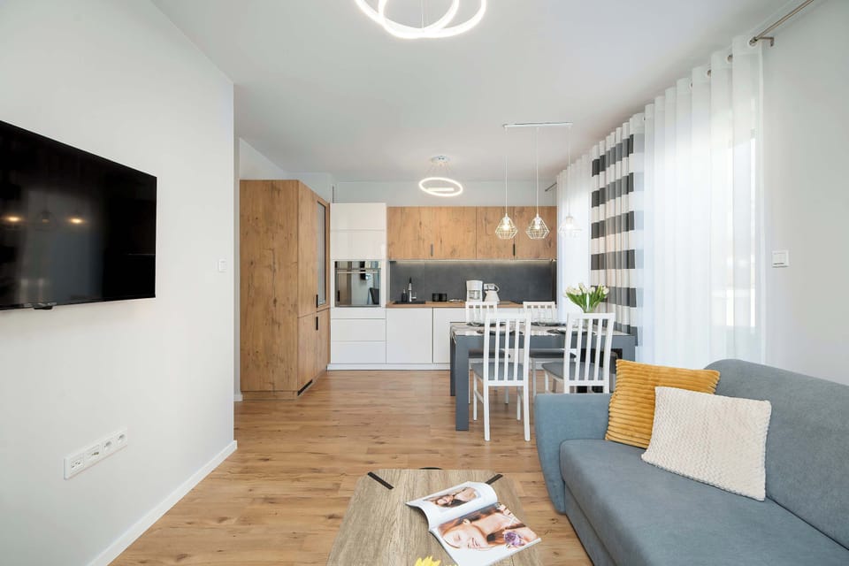 A living room featuring a wall-mounted television and a wooden media cabinet. A comfortable gray sofa faces the TV, creating a relaxing entertainment space. The room has a minimalist design with light walls and wooden flooring.