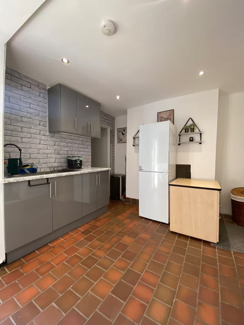 The kitchen viewed from the doorway end, showing grey cabinetry, a white fridge freezer, and terracotta-style floor tiles.