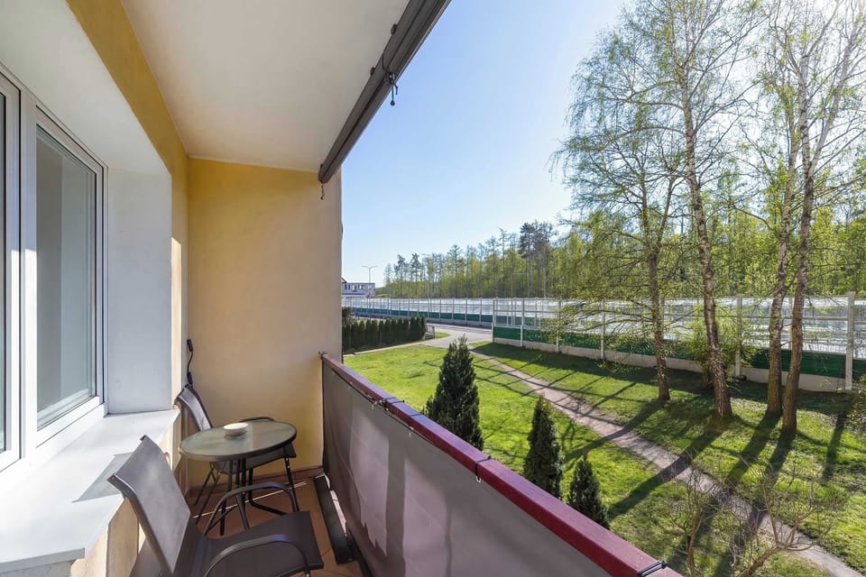 A balcony with a railing overlooking green trees and an open outdoor area. A small chair and table create a quiet place to sit and enjoy the view. The long corridor-like balcony stretches along the building exterior.