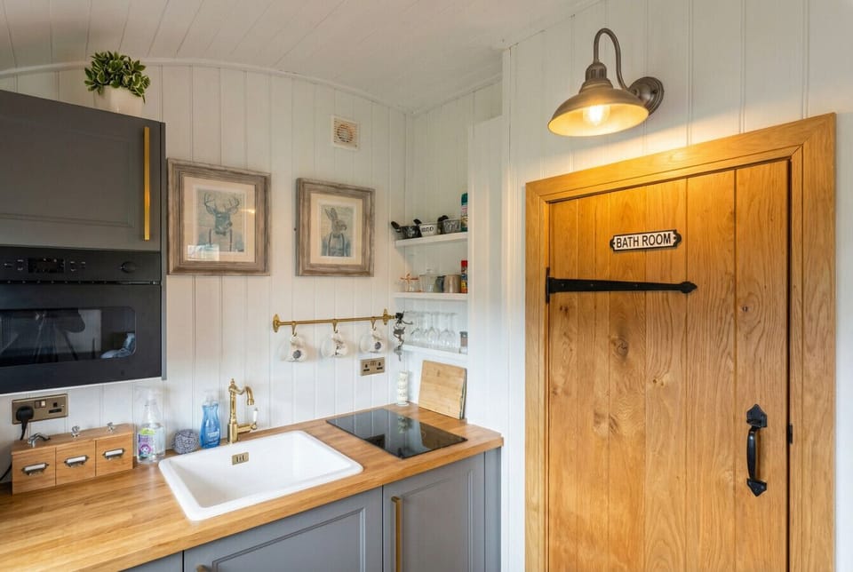 Cottage-style kitchen flows through a charming oak bathroom door lit by a vintage wall lamp.