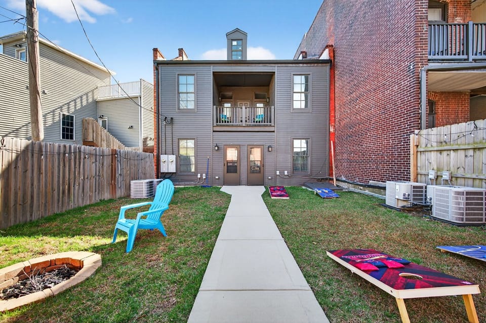Outdoor Entertainment: Step outside for fun and relaxation in this fenced backyard space featuring a central fire pit, cornhole boards for lawn games, colorful outdoor seating, and a paved walkway leading directly from the building.