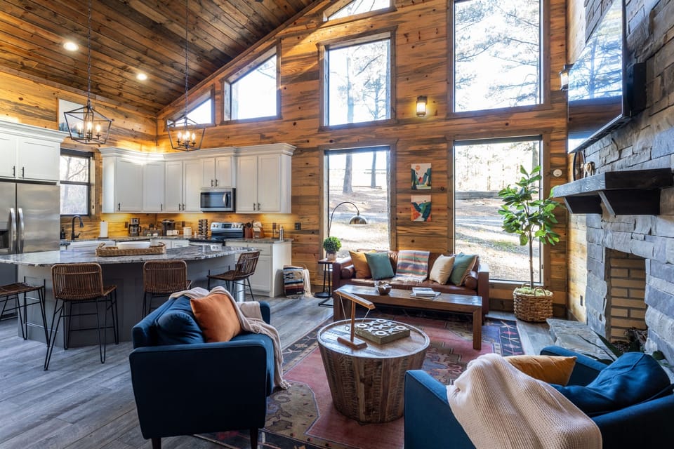 This living space features large windows for natural light. The open room integrates a modern kitchen with a lounge area, featuring plush blue chairs, a leather sofa, a gas fireplace, and a large smart TV.