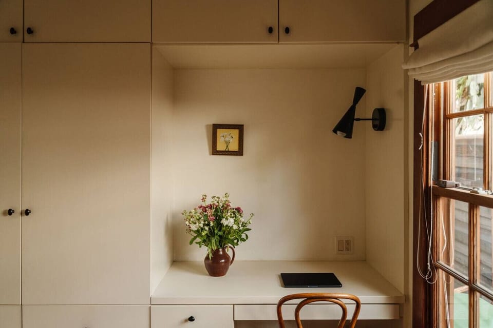 Warm, minimal bedroom with a reading nook, soft sconce lighting, fresh florals, and a wood-framed window — simple and serene.