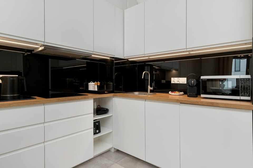 A modern kitchenette with sleek cabinets and built-in appliances. White countertops and walls create a clean contrast with the dark cabinetry. The space is compact but fully functional for preparing meals.