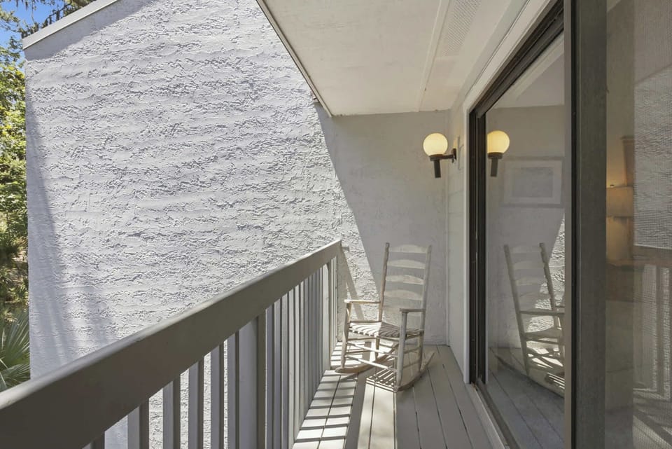 Weathered wood and soft shadows transform this private balcony into a peaceful morning nook. A classic rocking chair sits by the glass door, catching the gentle outdoor brightness for a quiet moment of reflection.
