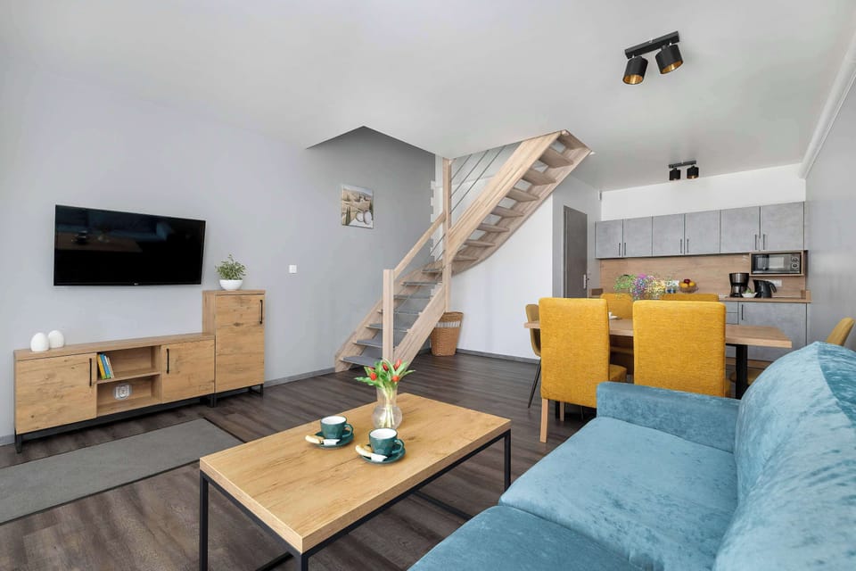 The living room view captures the wooden stairs and the entry to the kitchen area. A coffee table with cups is set in front of the sofa, making the space feel lived-in.