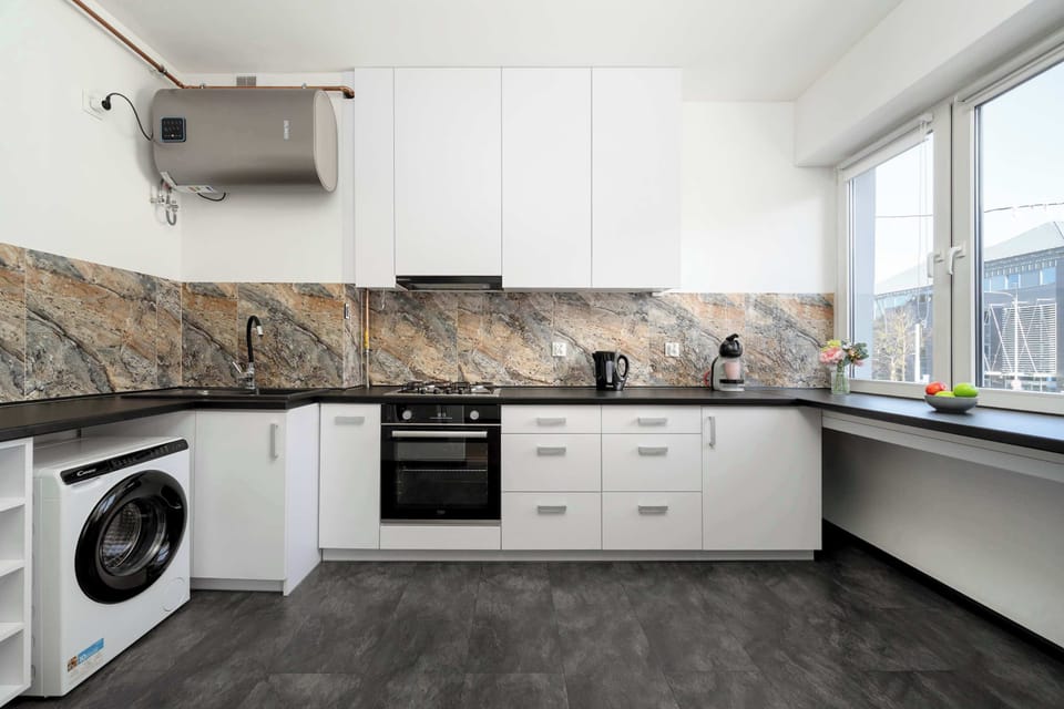 A wide view of a modern kitchenette with white cabinetry and a stone-pattern backsplash. Appliances such as a stove and washing machine are integrated into the lower cabinets. Large windows brighten the room with natural daylight.