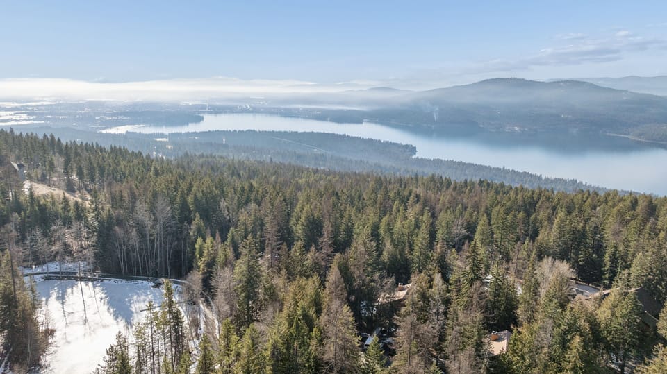 Stunning aerial perspective showcases the property's pristine winter setting among snow-dusted forests overlooking a serene lake and rolling mountains.