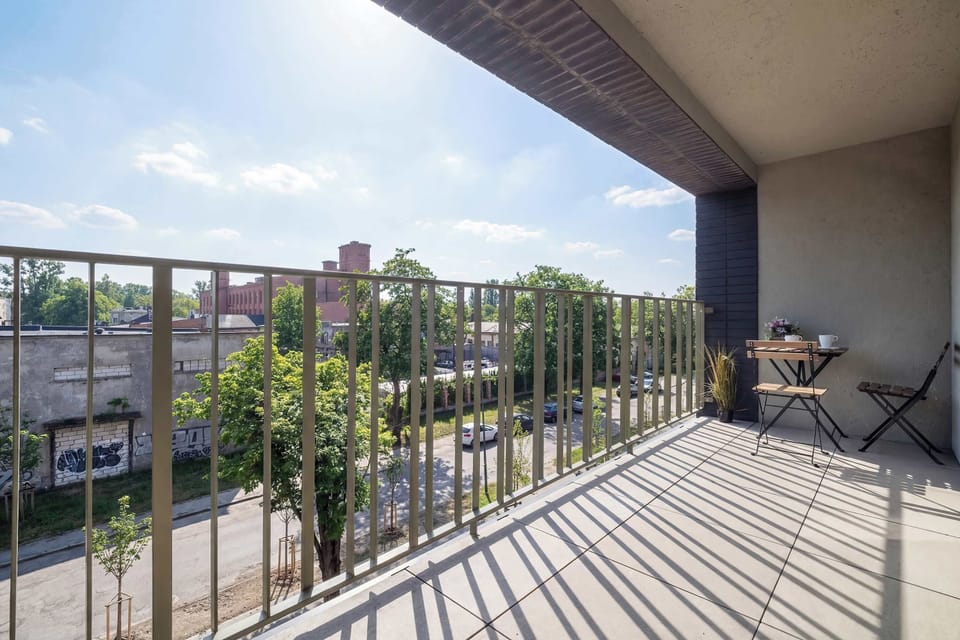 A long balcony with metal railings overlooking nearby buildings and trees. The outdoor area appears spacious enough for relaxing or enjoying the view.