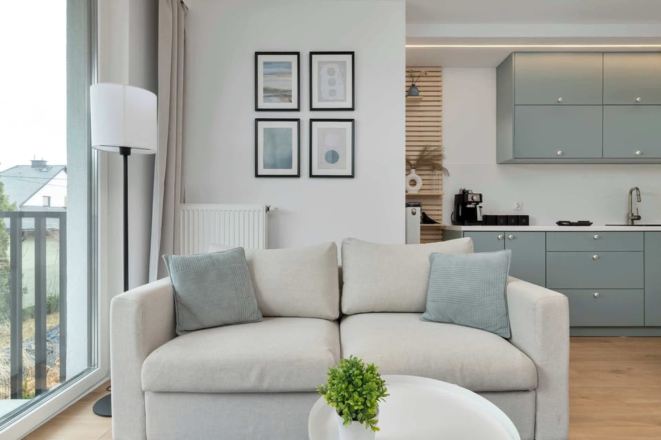 A comfortable sofa positioned near tall windows with a view, accompanied by a small plant on a round side table. The space connects visually to the kitchen behind it.