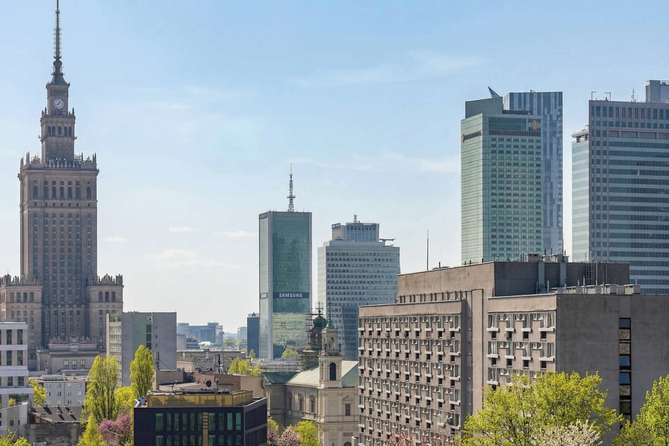 A stunning urban panorama captures the Warsaw skyline, dominated by the iconic spire of the Palace of Culture and Science. The view stretches across various modern skyscrapers and historical buildings under a clear blue sky.