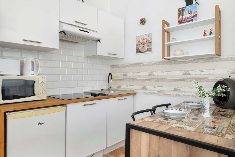 A minimalist kitchen corner includes sleek white cabinetry, a built-in microwave, and a small refrigerator. Adjacent to it, a portion of the wooden dining table is visible with a simple glass vase.