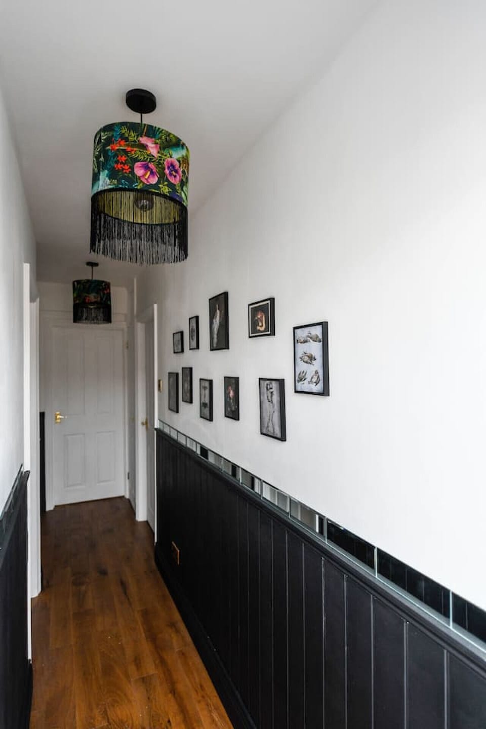 The hallway corridor is lined with tongue-and-groove wall panelling, a gallery of framed prints along one side, wooden flooring, and a decorative fringed lampshade.