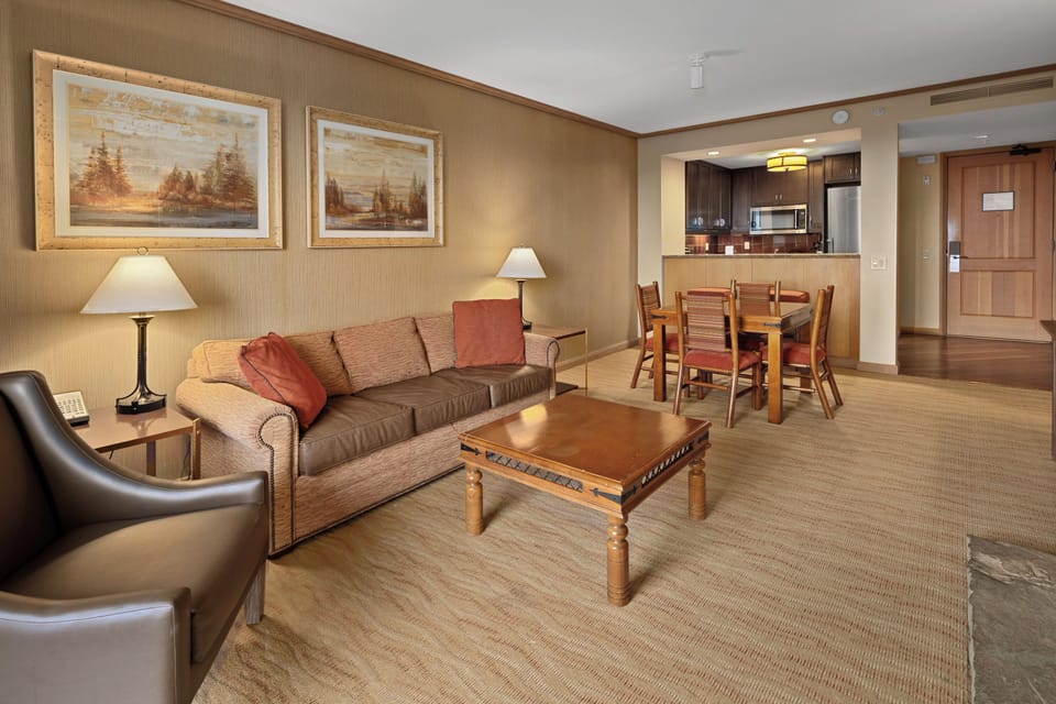 An open-concept suite living space with a sofa, armchair, coffee table, dining area for four, and a kitchenette with stainless steel appliances. Warm lighting and natural tones create a welcoming atmosphere.