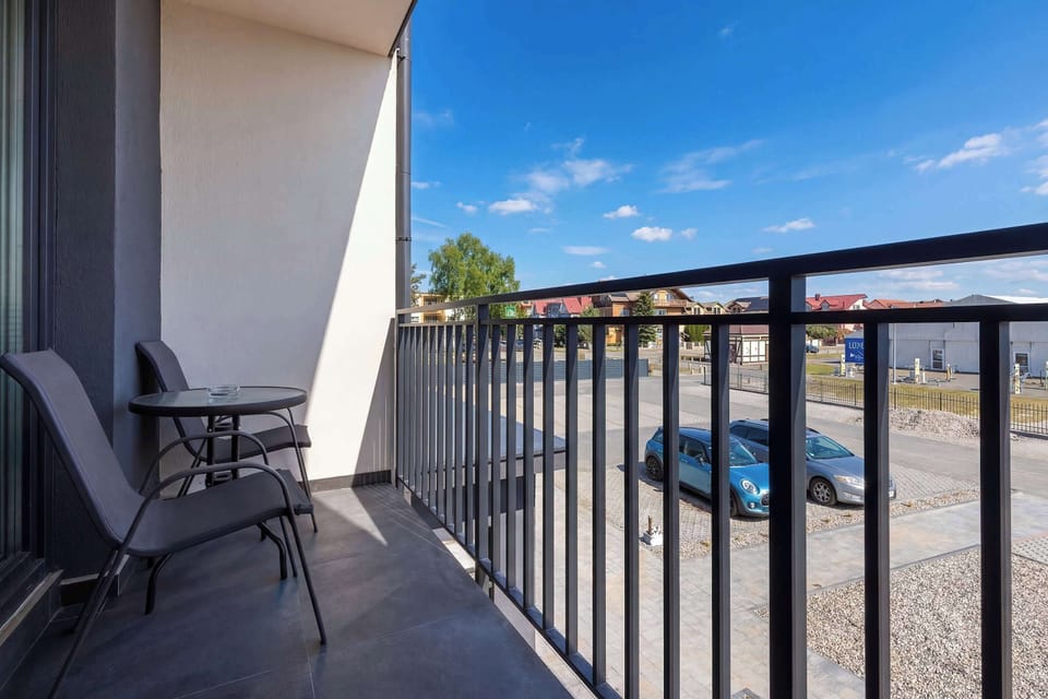 A balcony with a small seating setup and a view of the outdoors. Metal railings frame the edge, and the sky is clear. It looks like a peaceful spot to relax.