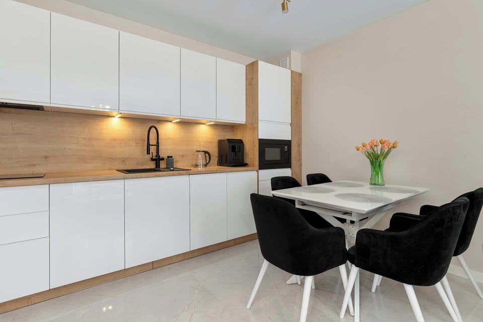 A dining space integrated with a sleek kitchenettte. Dark chairs surround a small dining table, contrasting with the white cabinetry. Under-cabinet lighting highlights the clean design.
