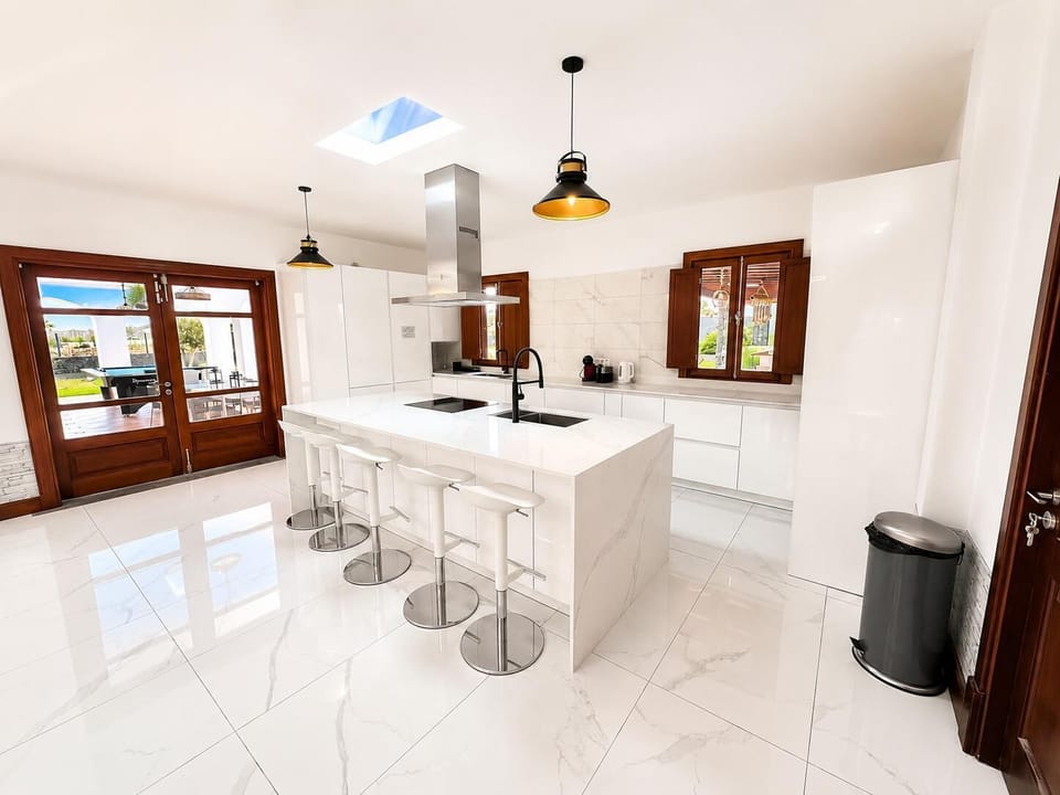 A sleek, modern white kitchen features a spacious waterfall island with barstools, high-end stainless steel appliances, and polished marble-style tile flooring.