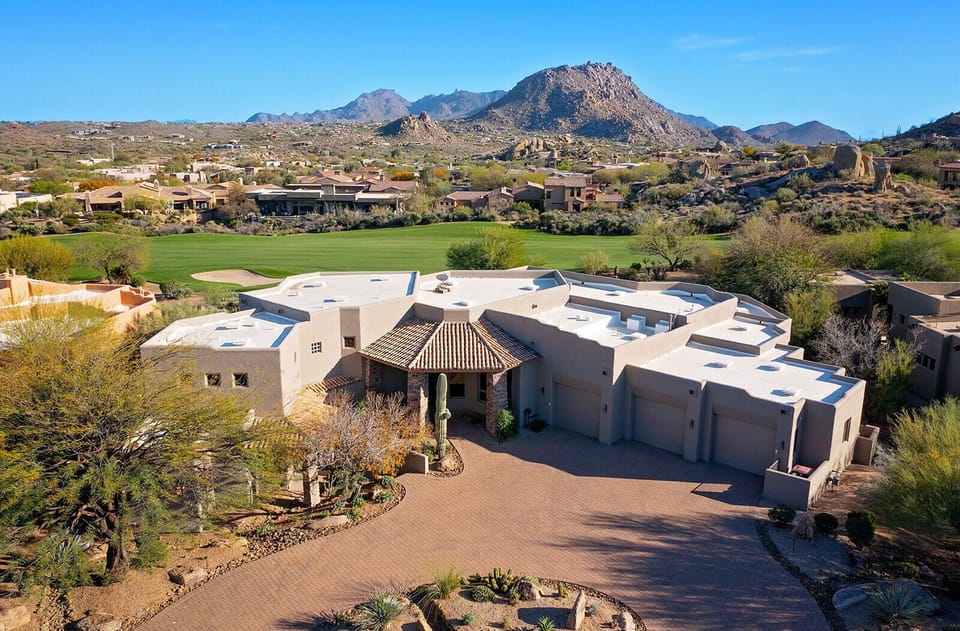 Aerial view of the home set against the desert hillside, with the golf course stretching out and rugged mountains rising in the distance. The curved driveway, natural desert landscaping, and quiet surroundings make it feel private and tucked away.