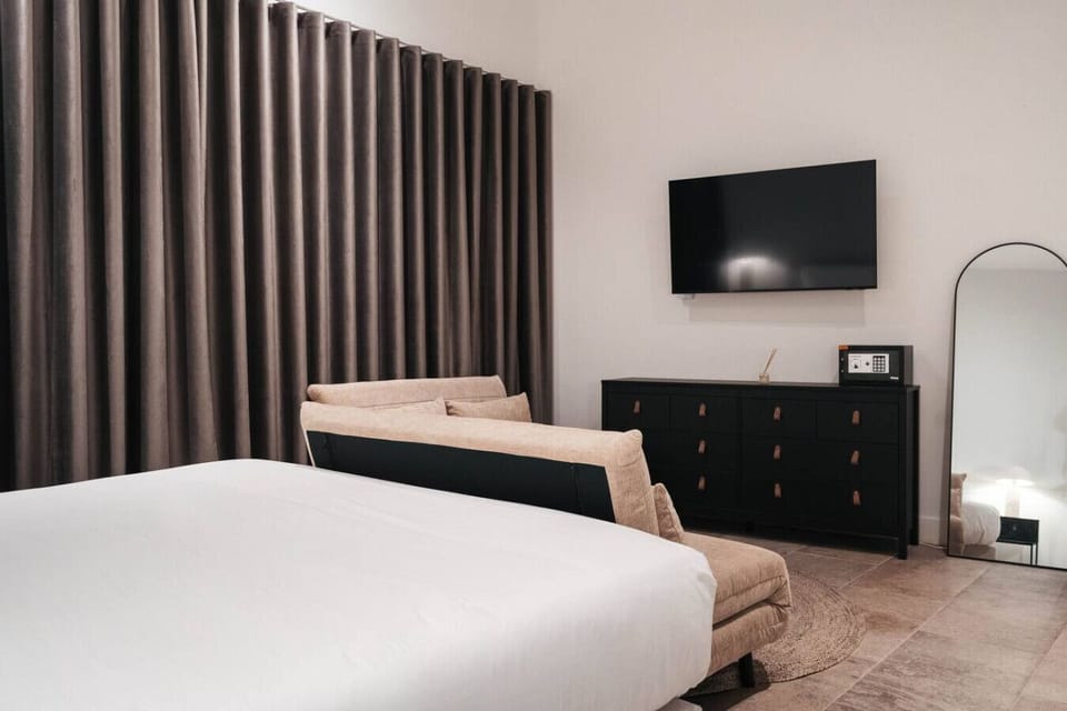 Relax in a calm, modern bedroom with a plush king bed, cozy lounge seating, and a wall-mounted TV—perfect for unwinding after a day out.