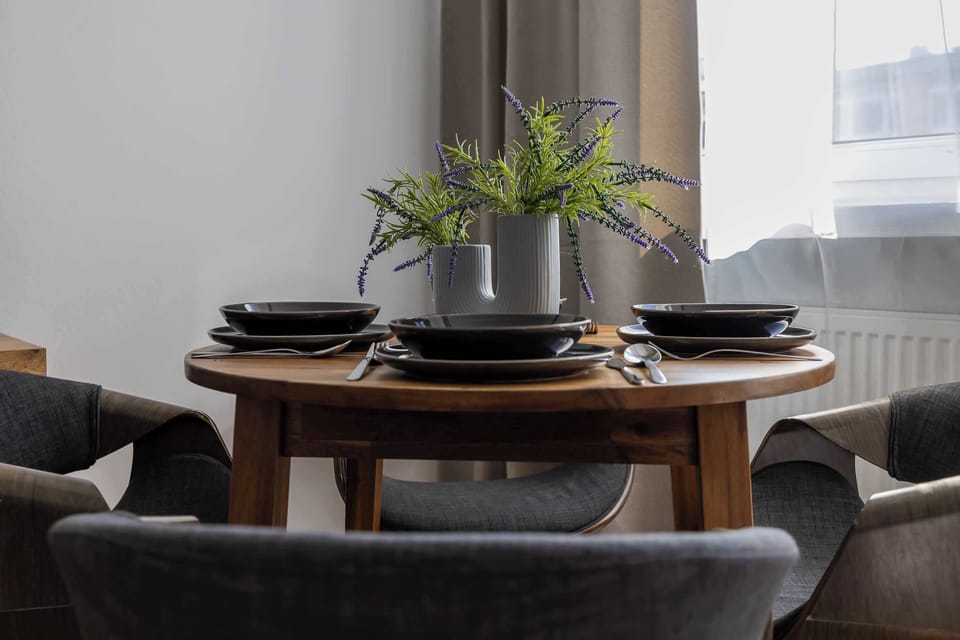 The round wooden table in the dining area has been carefully set with black tableware for four, highlighting the apartment’s readiness to welcome guests. Fresh plants in a vase add a natural touch to the interior and enliven the space.
