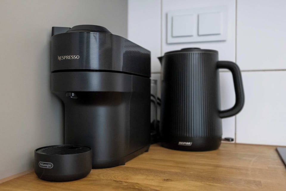 A high-quality black electric kettle and a coffee maker are placed on the kitchen countertop, promising enjoyable mornings. The stylish black accessories blend perfectly with the wooden countertop and white brick on the wall.