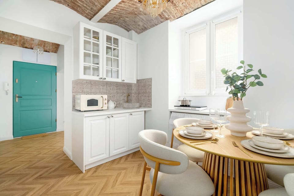 The kitchen area blends modern white cabinetry with rustic charm, highlighted by a vibrant turquoise door. A small dining setup sits near a window, while checkered wooden floors tie the natural textures together.
