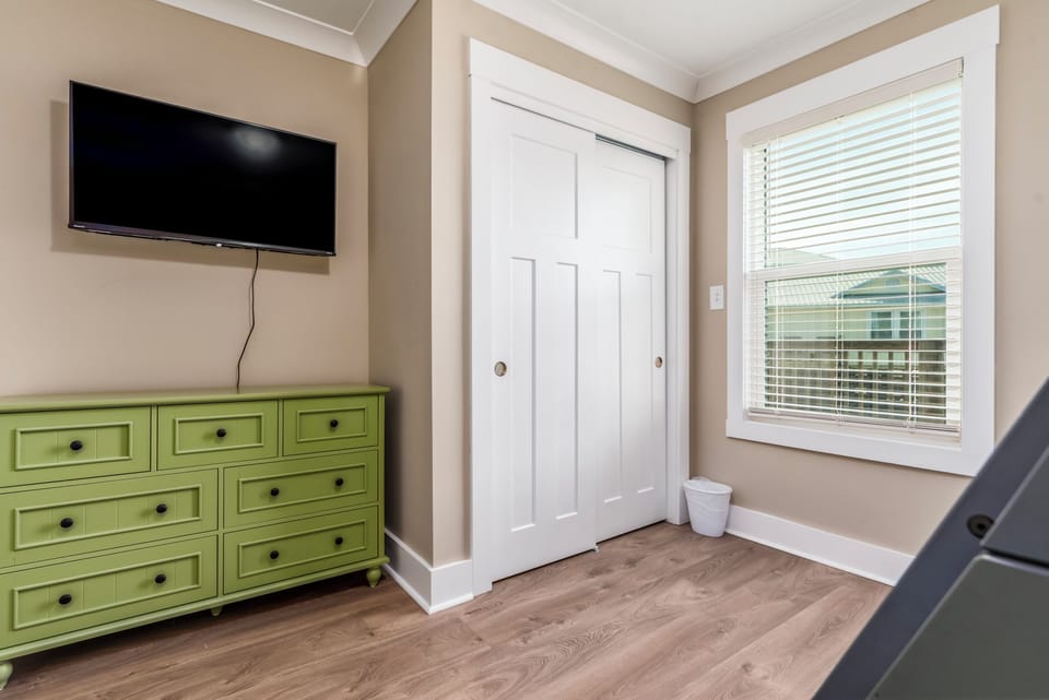 Your peaceful retreat features a wall-mounted TV and charming green dresser, with natural light streaming through blinds to create a cozy atmosphere.