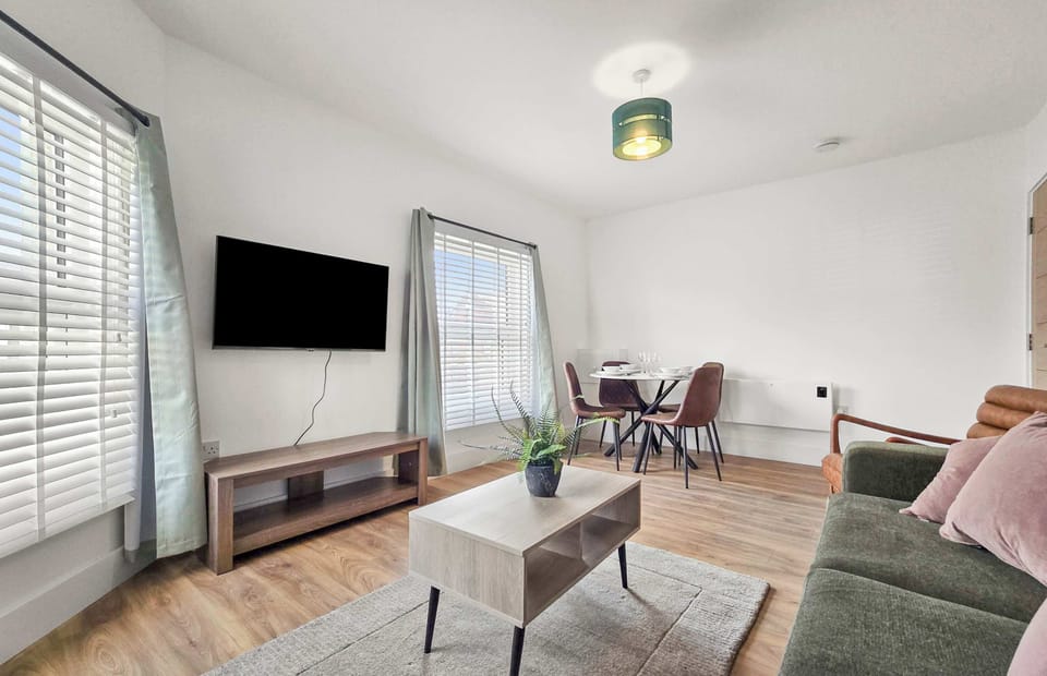 A bright, modern living room featuring minimalist furniture, wooden floors, and a wall-mounted TV. Large windows fill the space with natural light, illuminating both the seating and dining areas.
