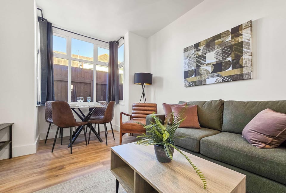 This living room features a modern aesthetic with a green sofa, a light wood coffee table, and a small dining area by the window. Earthy tones and natural light create a warm, inviting atmosphere in the space.