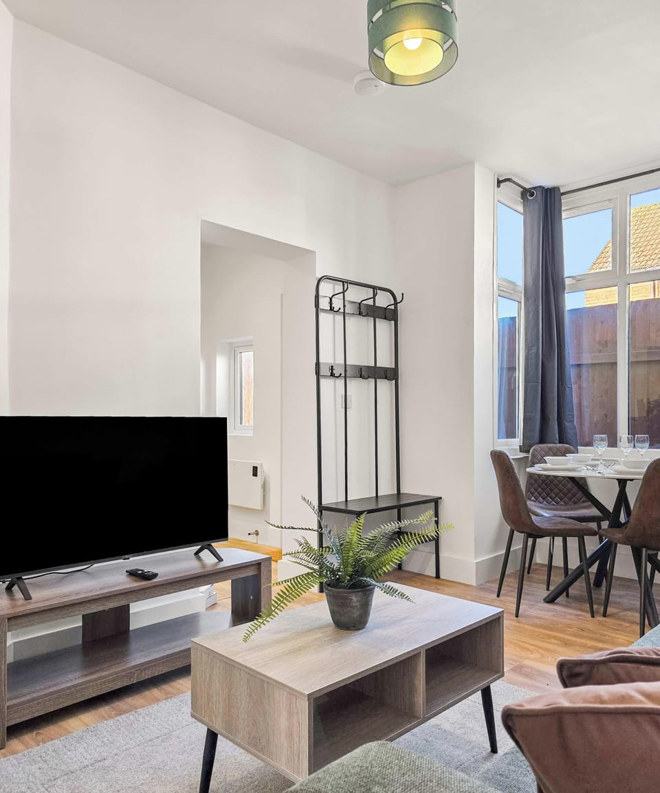  A brightly lit, modern living area featuring light wood furniture and a minimalist black coat rack. A large window with grey curtains illuminates a small dining set in the background.