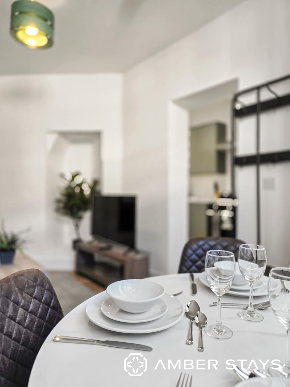 A shallow-depth-of-field shot showcases a formally set dining table with white dinnerware and glassware in the foreground. The background softly blurs into a modern living area featuring a television, indoor plants, and a peek into an adjacent room.
