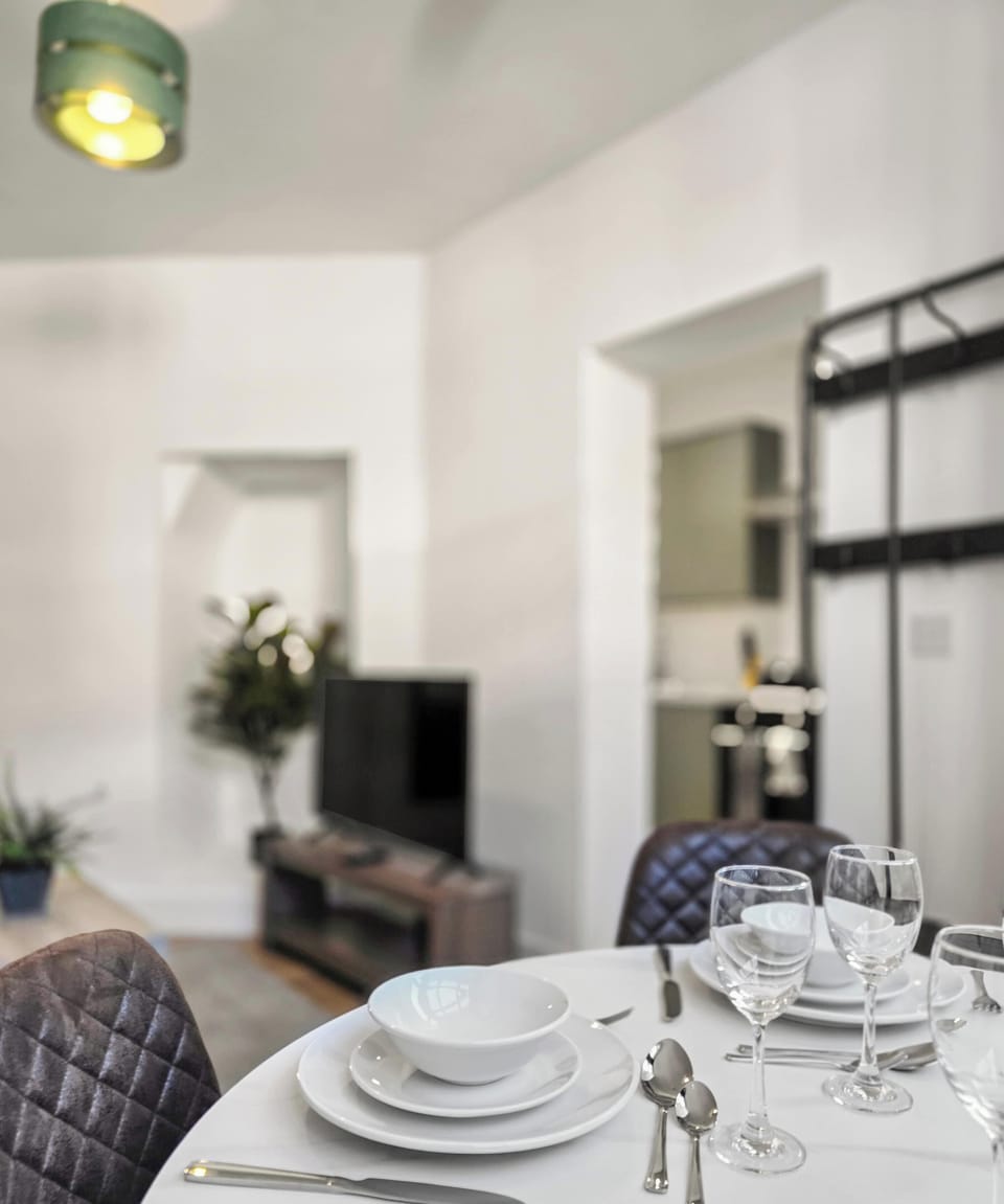 A shallow-depth-of-field shot showcases a formally set dining table with white dinnerware and glassware in the foreground. The background softly blurs into a modern living area featuring a television, indoor plants, and a peek into an adjacent room.
