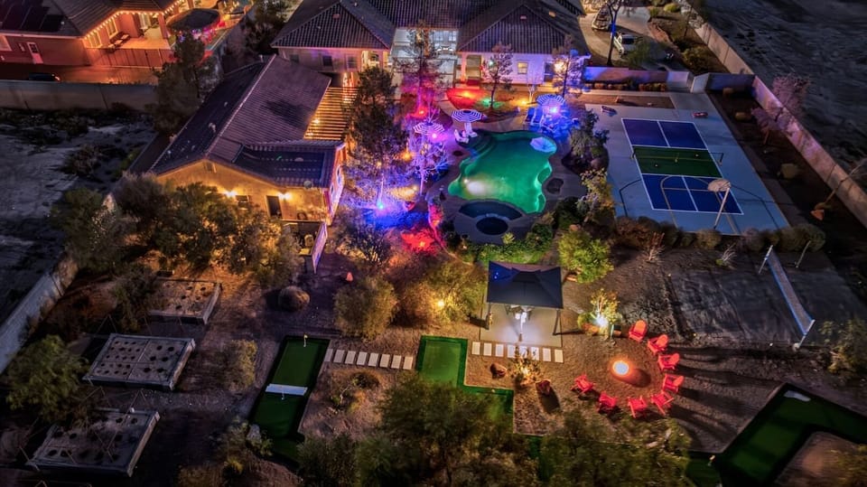 Stunning aerial view of the backyard at night featuring the pool and hot tub, pickleball and basketball court, mini golf course, fire pit lounge, and beautifully lit resort style grounds.