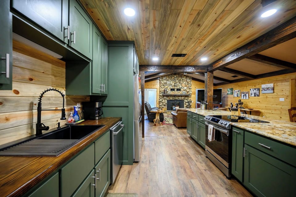 Your rustic chef's dream features sage green cabinets, granite countertops, and exposed beam ceilings that perfectly frame the cozy stone fireplace beyond.
