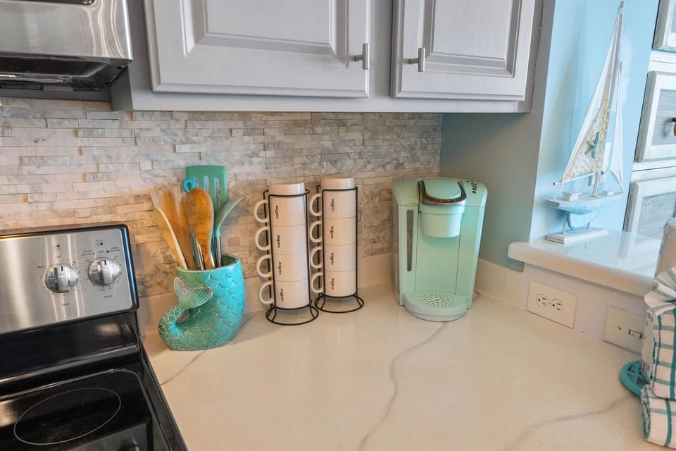 Your morning coffee station awaits — Keurig, stacked mugs, and coastal-charmed countertops with a view of the Gulf just steps away.