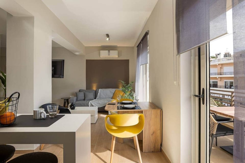 Bright open-plan living area with a comfortable sofa, wall-mounted TV, and a compact dining table. Large balcony doors bring in natural light and connect the room to an outdoor seating area, adding an easy indoor-outdoor feel.