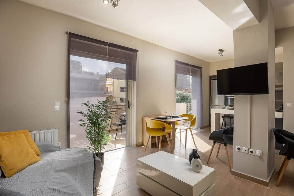 Bright open-plan living area with a comfortable sofa, wall-mounted TV, and a compact dining table. Large balcony doors bring in natural light and connect the room to an outdoor seating area, adding an easy indoor-outdoor feel.