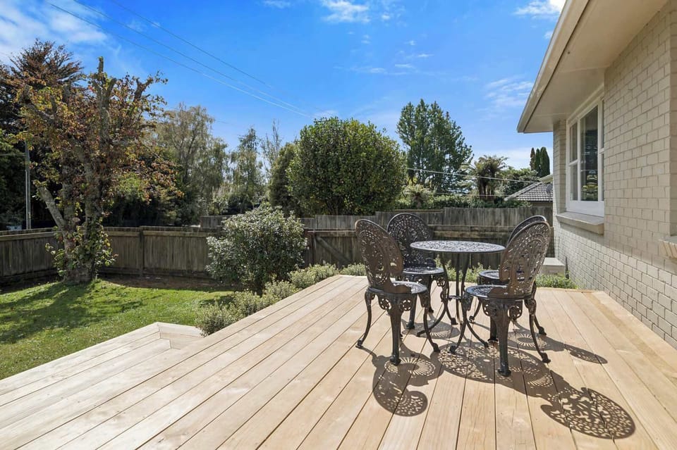 Front timber deck with wrought-iron bistro table and 4 chairs, raised garden beds, and steps to entry — sunny outdoor living at Red Door 56 Rotorua