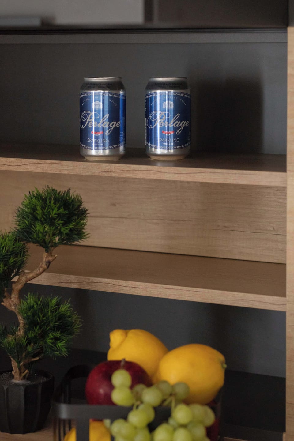 A detailed shot of an open kitchen cabinet reveals a small potted plant and a bowl of fresh fruit, including lemons and grapes. Above, two canisters are neatly stored on a wooden shelf.