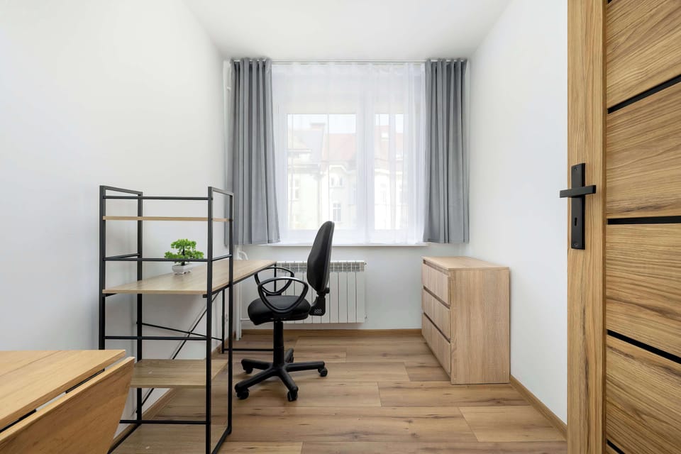 A dedicated workspace includes a modern desk with a black mesh office chair and integrated shelving. A large window provides plenty of light for working, and a wooden dresser sits nearby for storage.