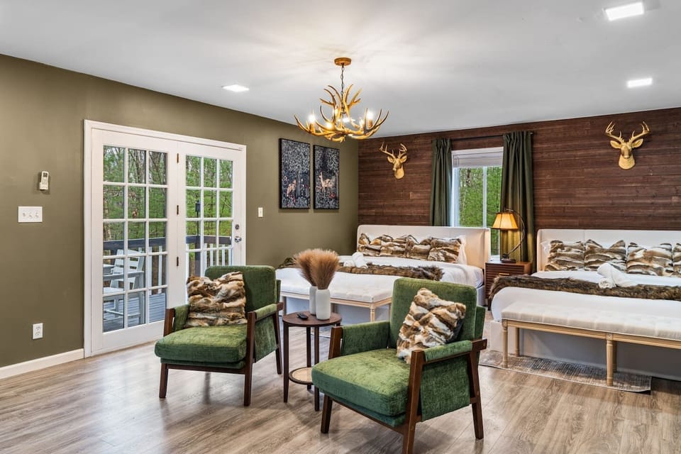Cozy, lodge inspired bedroom with two beds, warm wood accents, and a unique antler chandelier. French doors open to the deck, with a seating area perfect for relaxing and enjoying peaceful forest views.