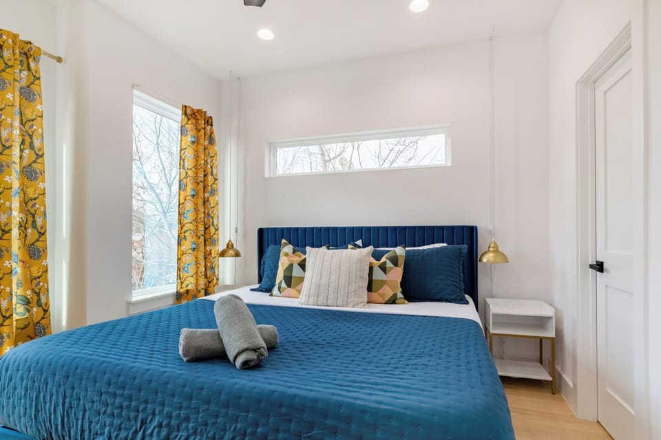This vibrant bedroom features a king-sized bed with a royal blue tufted headboard and matching linens, beautifully accented by gold-patterned drapery and modern bedside lighting.