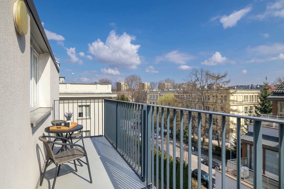 A private city-view balcony is furnished with a small table and chairs for two. The metal railing overlooks a quiet urban street under a bright blue sky.