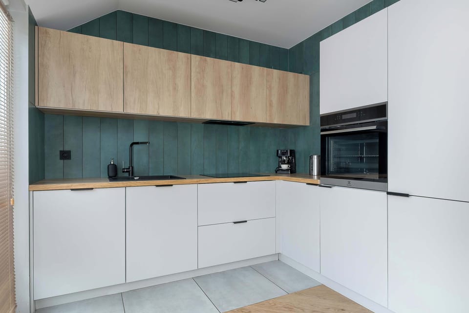 A sleek kitchenette layout displays white handleless lower cabinets contrasted with a dark teal backsplash and light wood upper units. The built-in oven and minimalist sink area maintain a clean, high-end look.