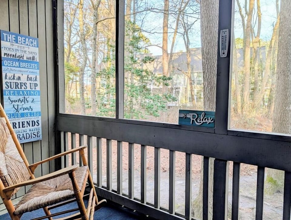 2306 Round Robin Way Sea Colony West Screened Porch with Rocking - A screened-in outdoor balcony porch featuring a wooden rocking chair, timber railings, and a view facing dense green woods.