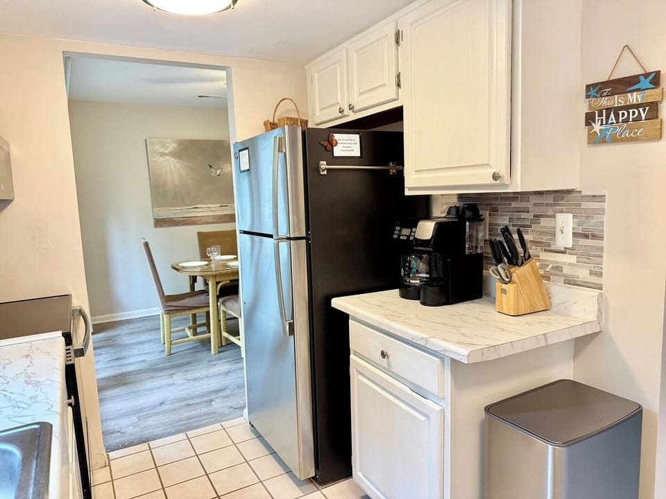 2306 Round Robin Way Sea Colony West Modern Kitchen White Cabine - A kitchen interior showcasing white cabinetry, a stainless steel refrigerator, white countertops with a coffee maker, and a view leading toward the dining area.